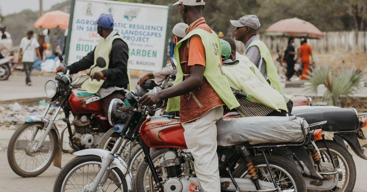 Group Of Motorbike Riders Wearing Vests In A Bustling African City Street Scene.